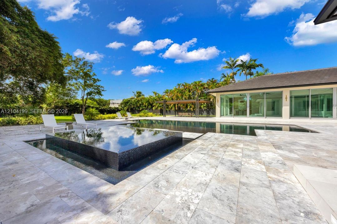 Backyard featuring a custom infinity-edge pool, a raised square spa with tile surfacing, extensive stone paver decking, and a covered pavilion structure