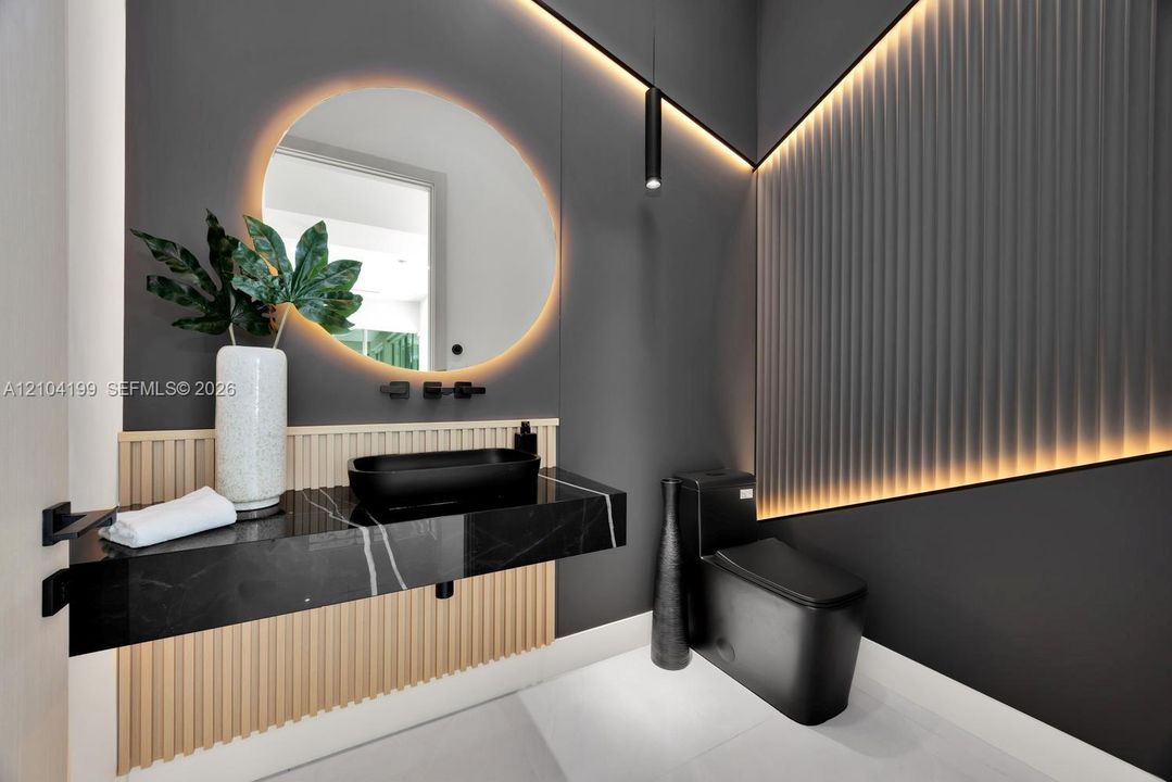 Floating vanity with stone countertop and matte black vessel sink, circular backlit mirror, vertical slat wood accent paneling, integrated recessed cove lighting, and matte black plumbing fixtures