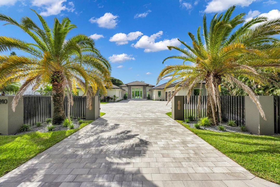 Gated entrance featuring a wide paver driveway, concrete pillar accents, vertical metal fencing, and mature palm landscaping