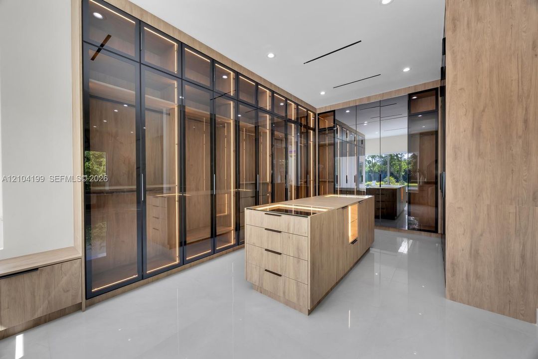 Custom storage system featuring floor-to-ceiling cabinetry with glass-panel doors, integrated LED lighting, central built-in island with storage drawers, wood-finish cabinetry, and high-gloss flooring