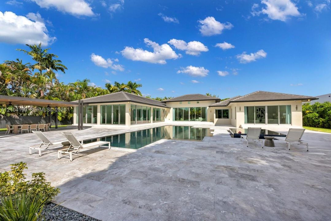 Spacious outdoor living area featuring extensive stone paver hardscaping, an in-ground swimming pool, a covered pavilion, and floor-to-ceiling glass panel doors