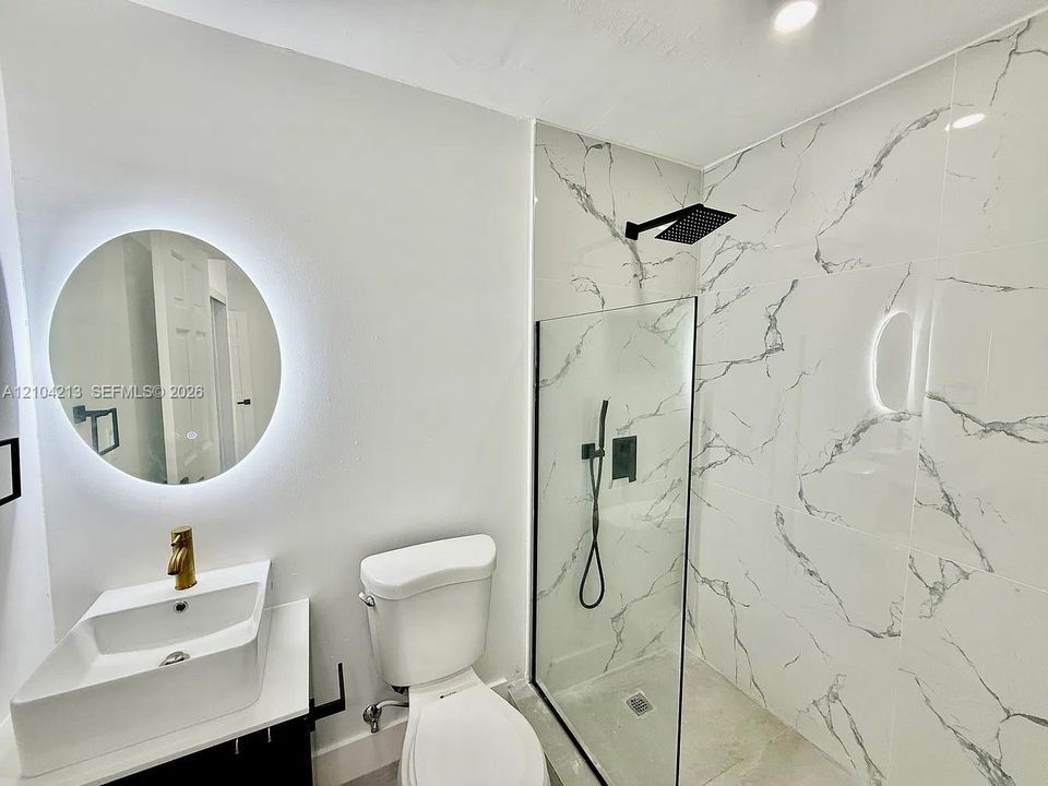 Modern bathroom featuring a rectangular vessel sink with gold-finish faucet, an illuminated circular mirror, and a walk-in shower with black-finish fixtures and patterned wall tile