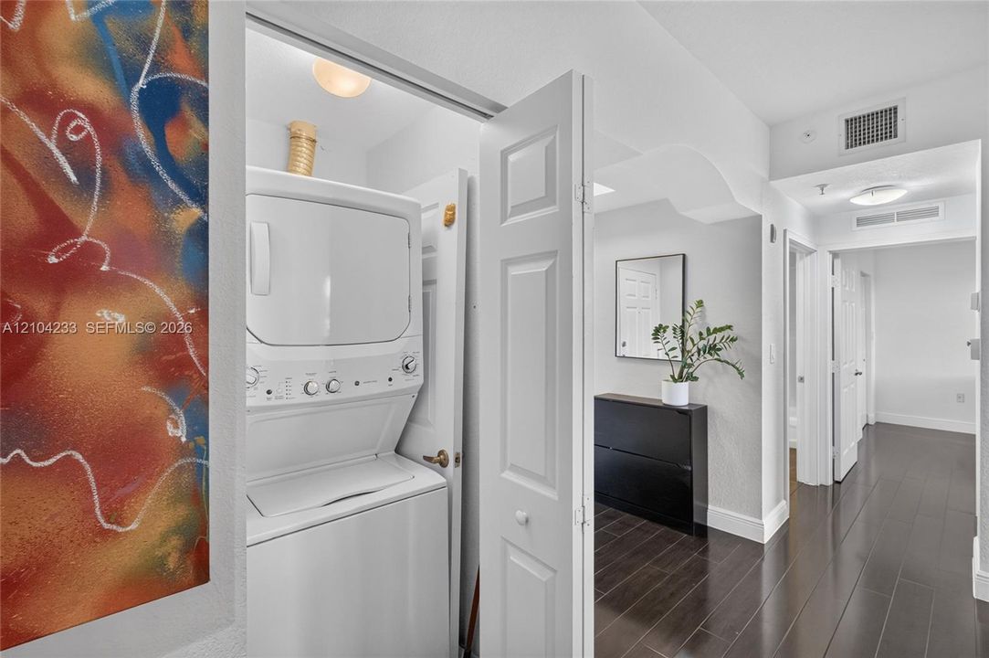 Laundry closet featuring a white stacked washer and dryer unit, recessed ceiling lighting, and wood-finish flooring throughout the hallway
