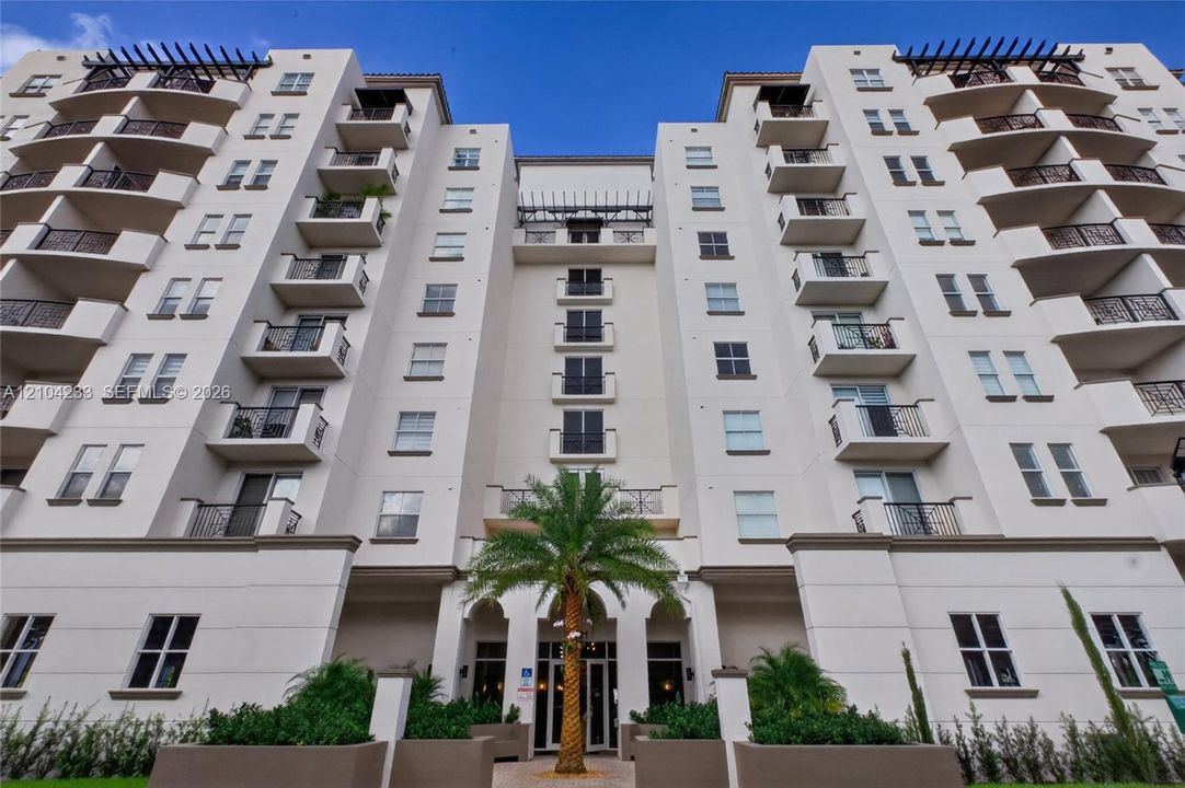 Multi-story stucco exterior featuring symmetrical design, private balconies with decorative metal railings, decorative roof pergolas, and palm-accented entrance hardscaping