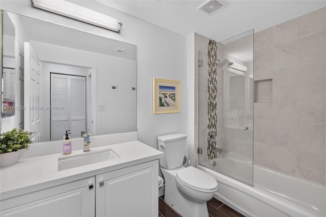 Bathroom featuring a vanity with a white countertop and rectangular undermount sink, a large wall-mounted mirror, a tub-shower combination with glass paneling, a vertical tile accent border, and recessed wall shelving