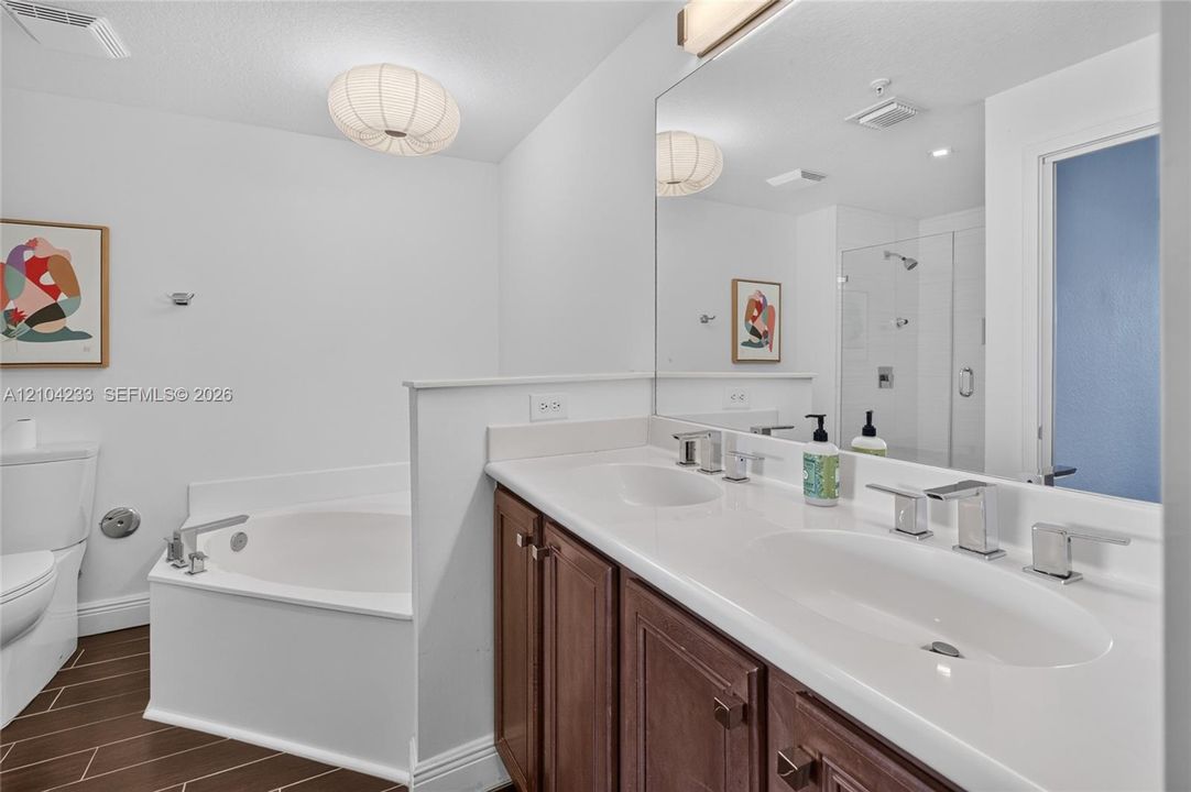 Bathroom featuring a dual-sink vanity with wood-finish cabinetry, a white countertop, a drop-in soaking tub, and a glass-enclosed shower with tile surround