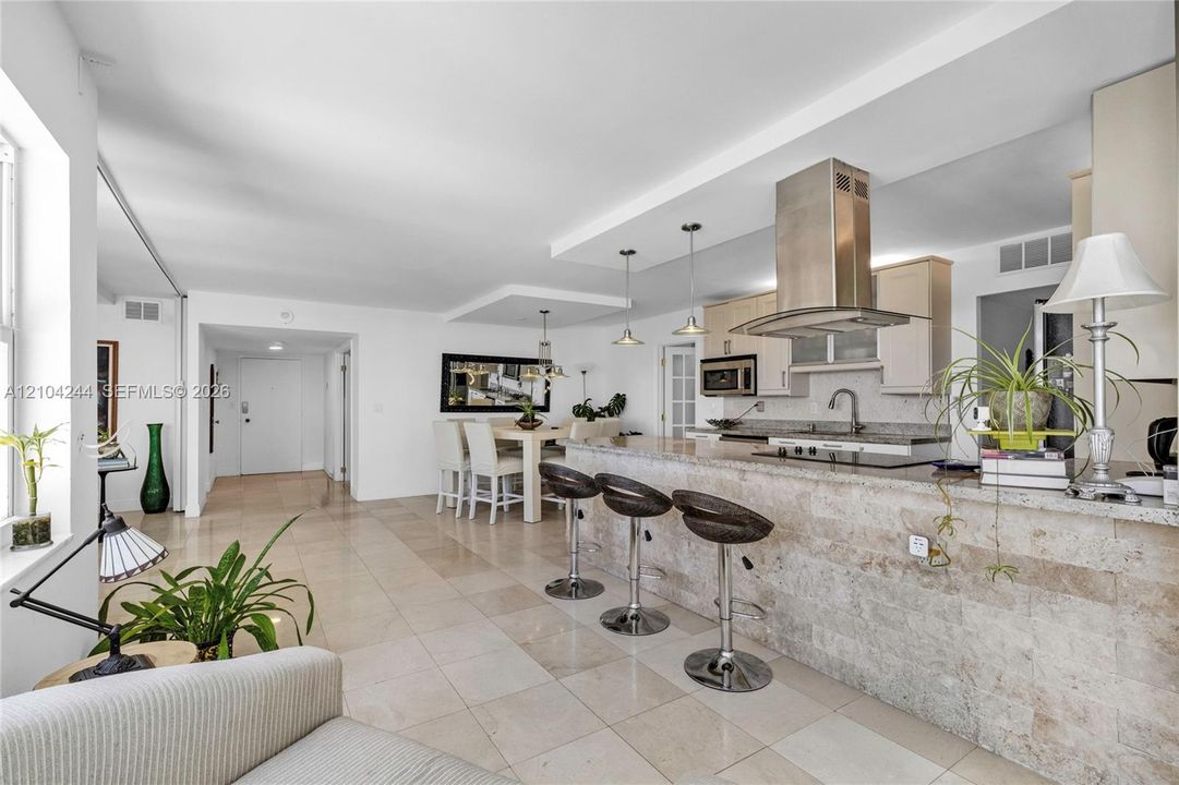 Open-concept kitchen featuring stone-clad breakfast bar, stainless steel chimney-style range hood, light-toned cabinetry, and tile flooring