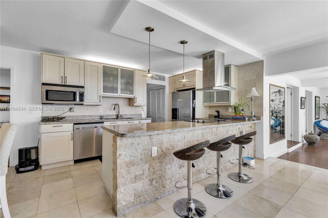 Kitchen featuring stone-clad breakfast bar, stone countertops, stainless steel appliances, cabinet-fronted upper storage with glass inserts, and two pendant lights suspended from a drop ceiling