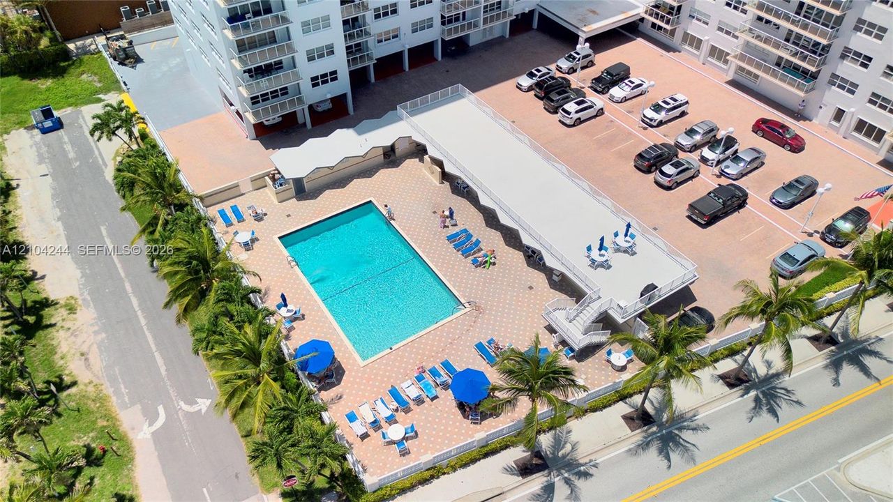 Aerial view of the building exterior featuring an in-ground swimming pool, paved pool deck, multi-level parking area, and landscaping with palm trees