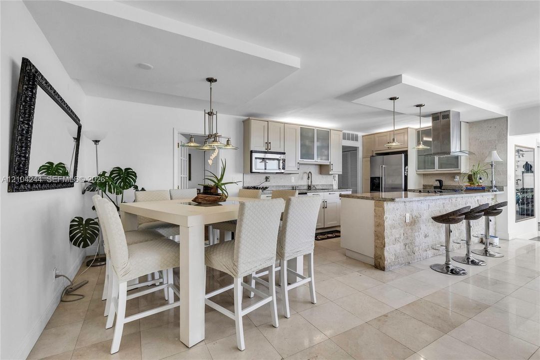 Open-concept kitchen featuring light wood cabinetry, stone-clad kitchen island, stainless steel appliances, tiered tray ceilings, and neutral tile flooring