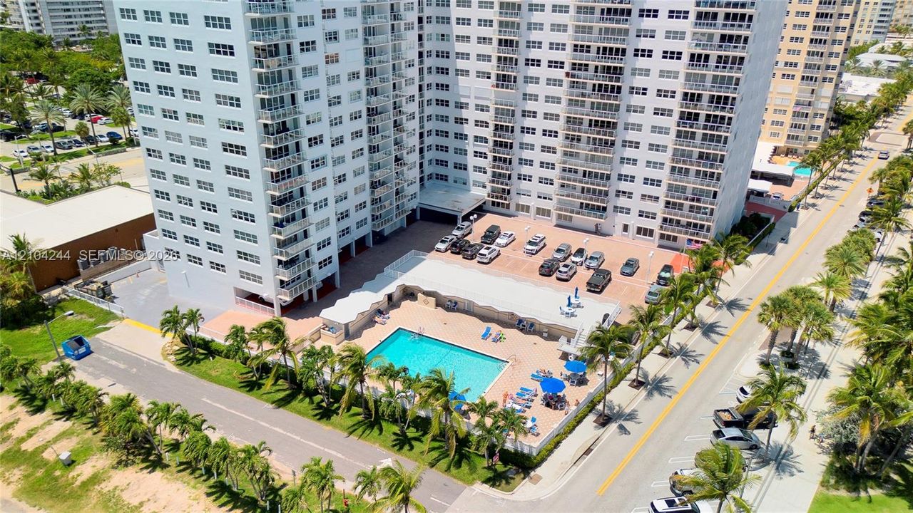 High-rise residential building featuring an outdoor swimming pool, paved surface parking, and perimeter landscaping with palm trees