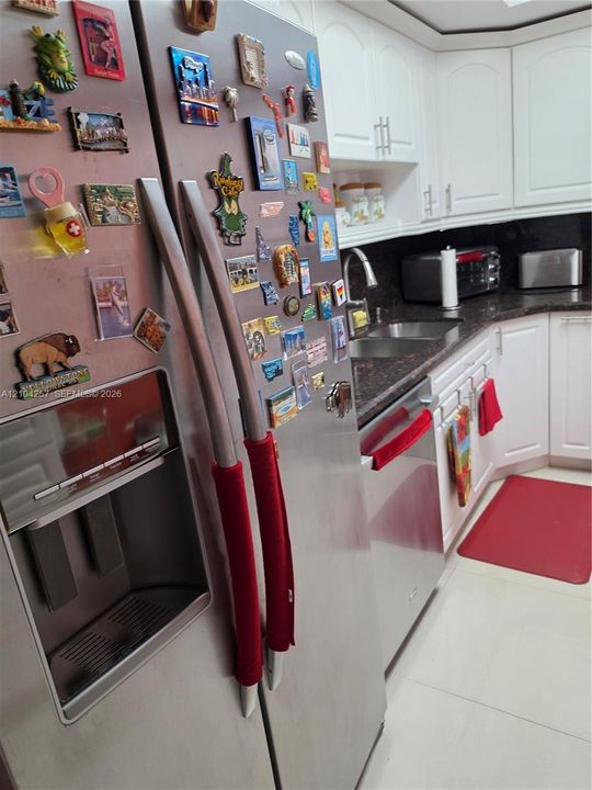 Stainless steel refrigerator with integrated water and ice dispenser, white shaker-style cabinetry, dark stone-finish countertops, dual-basin under-mount sink, and stainless steel dishwasher