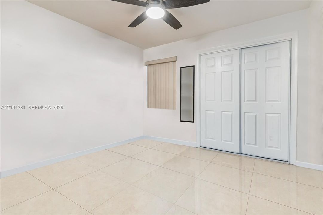 Neutral-toned tile flooring, white-panel sliding closet doors, flush-mounted ceiling fan with integrated lighting, and vertical window blinds
