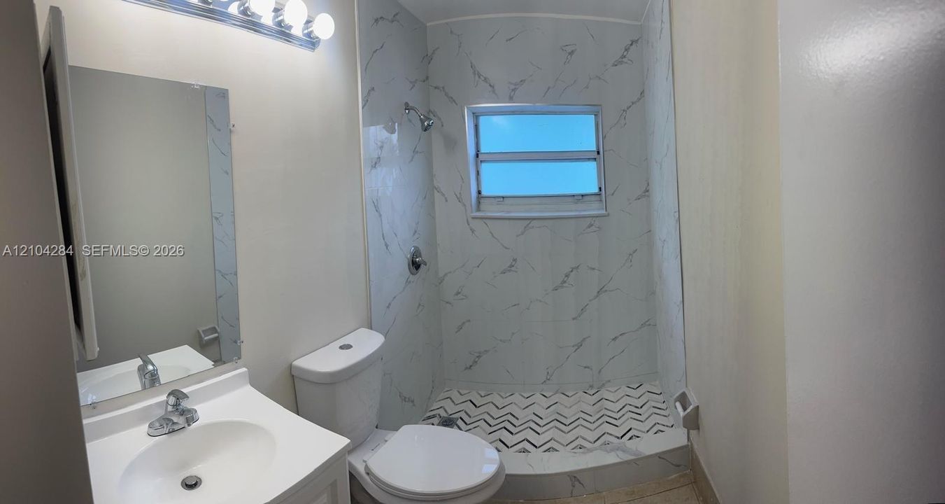 Bathroom featuring a walk-in shower with patterned floor tile, stone-finish wall surround, white pedestal vanity, and wall-mounted multi-bulb light fixture