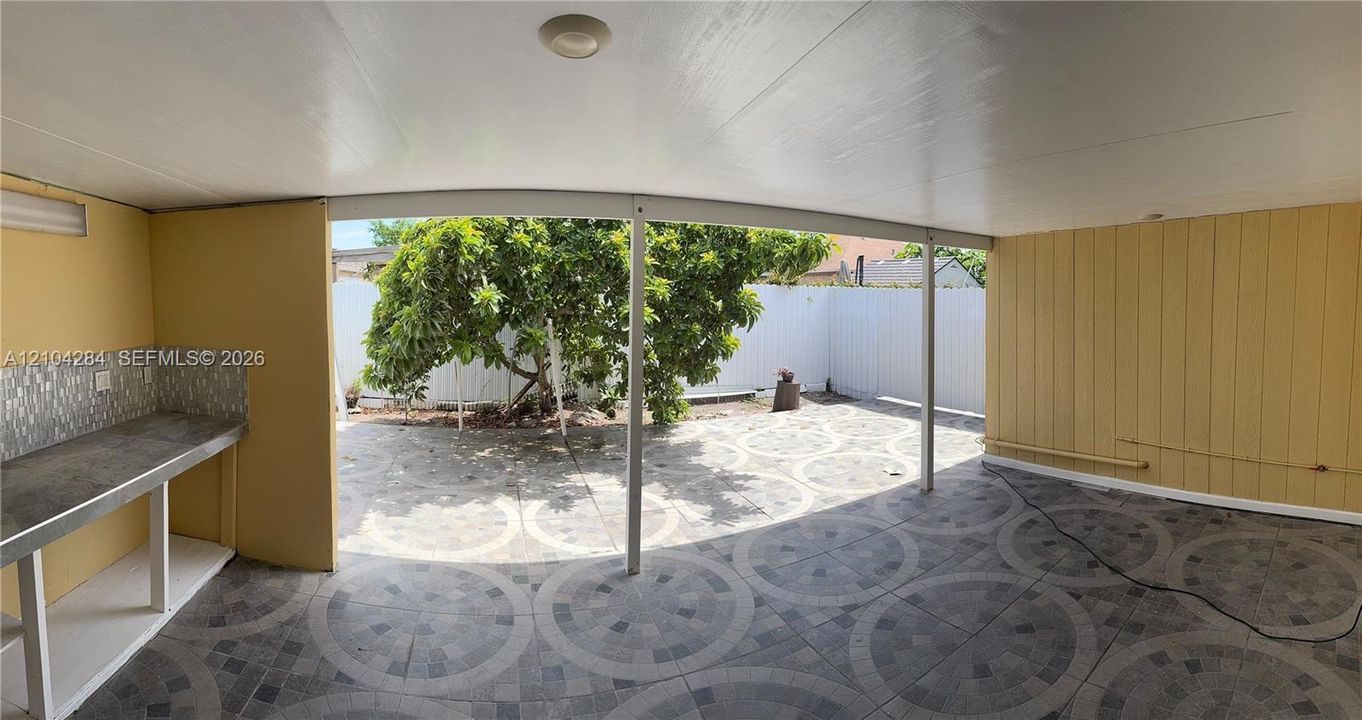 Covered patio featuring patterned tile flooring, built-in counter with mosaic tile backsplash, solid white privacy fencing, and mature tree