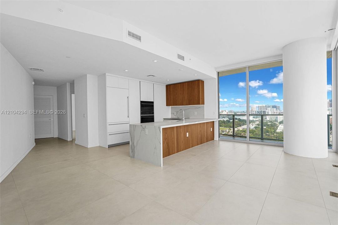 Open concept living area with light-toned tile flooring, floor-to-ceiling windows, and a modern kitchen featuring wood-finish cabinetry, integrated appliances, and a waterfall island