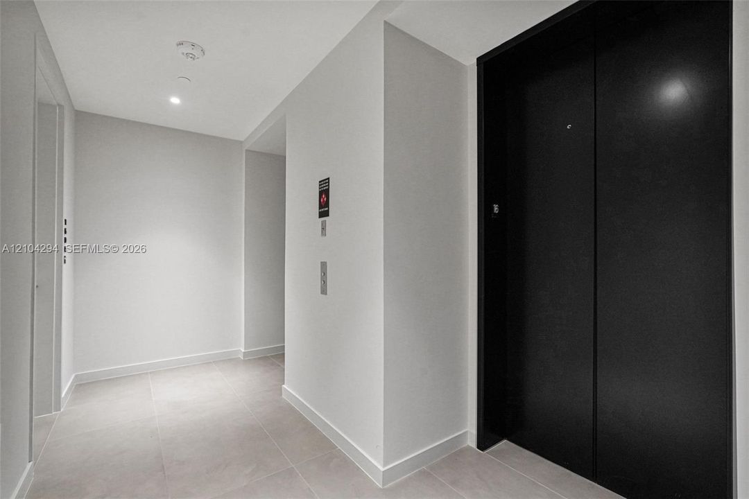 Recessed ceiling lighting illuminates the corridor, featuring a black elevator entrance, light-toned wall finishes, and square tile flooring
