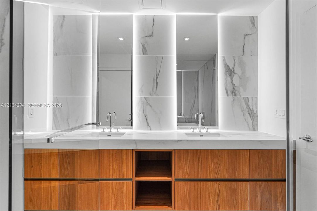 Dual vanity featuring wood-finish cabinetry, integrated sinks, a solid surface countertop, and backlit mirrors with wall-mounted stone-look tiles