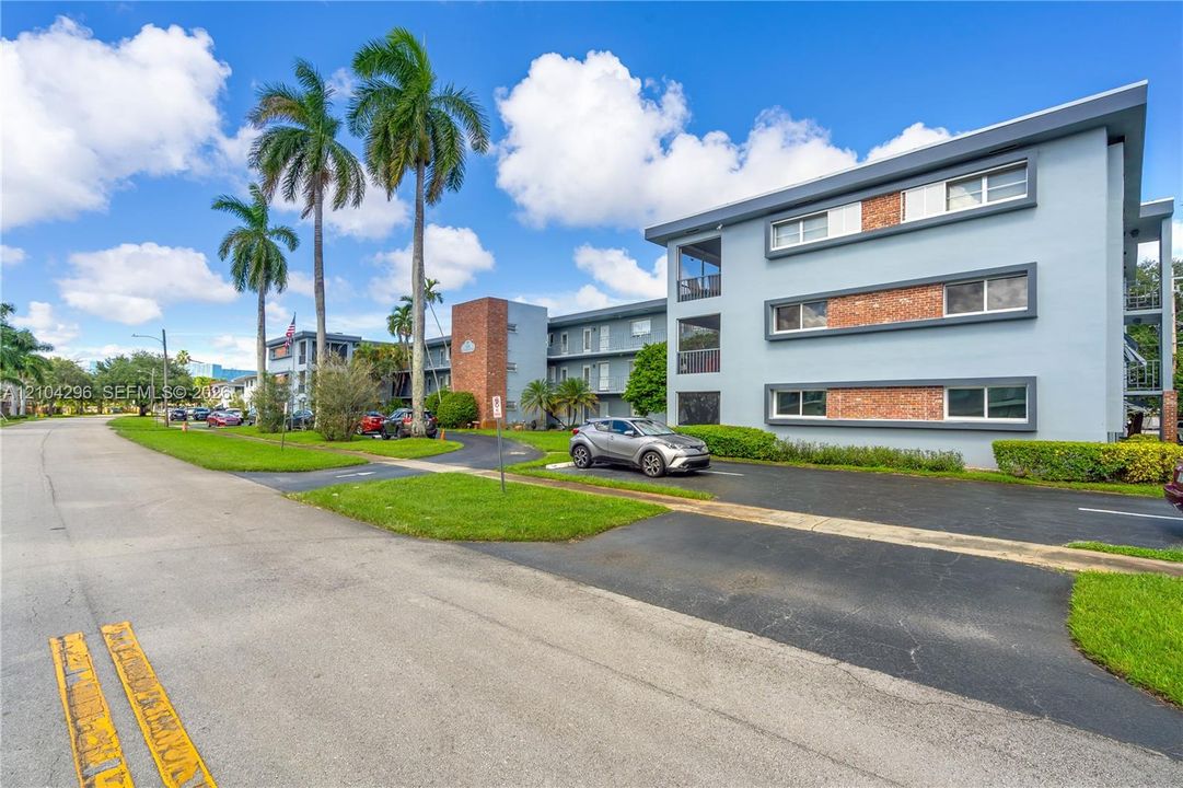 Three-story multi-family exterior featuring light-colored stucco finish, brick accents, rectangular windows with frames, and paved parking areas