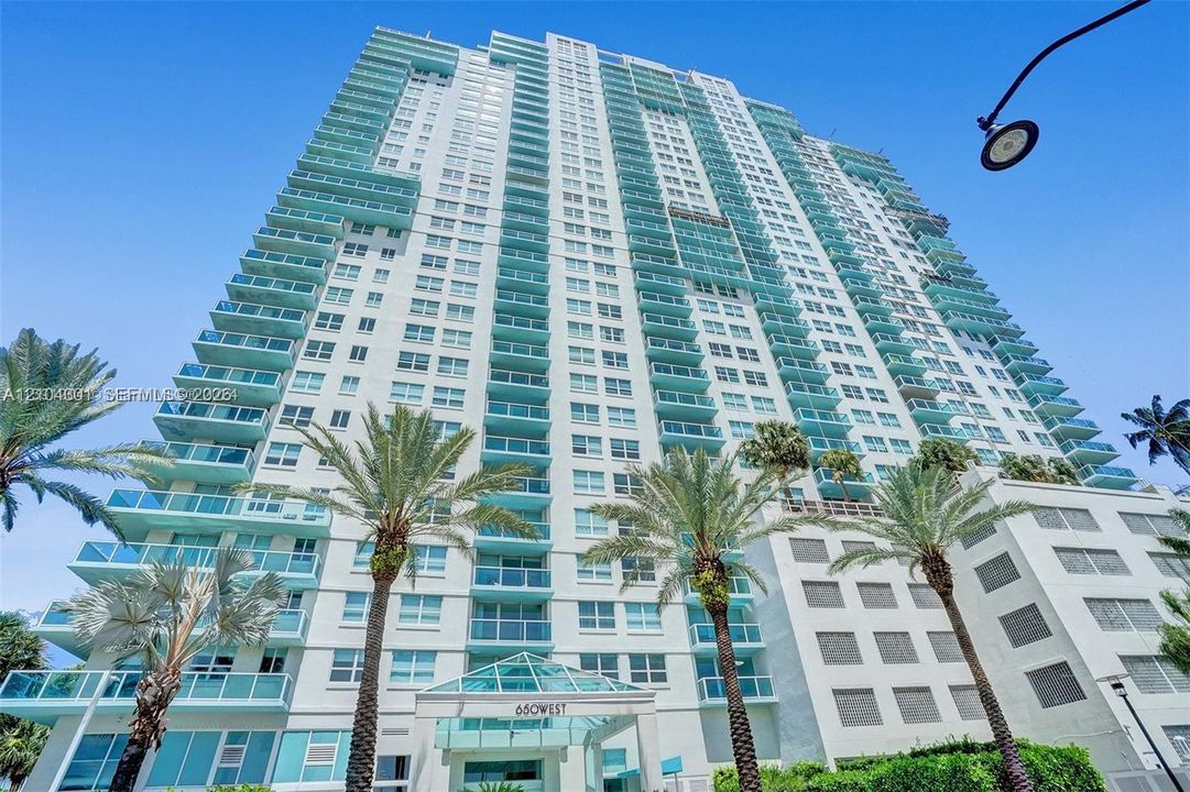 High-rise residential exterior featuring a light-toned facade, multiple levels of private glass-railed balconies, and a geometric glass-paneled entrance canopy, accented by mature palm trees