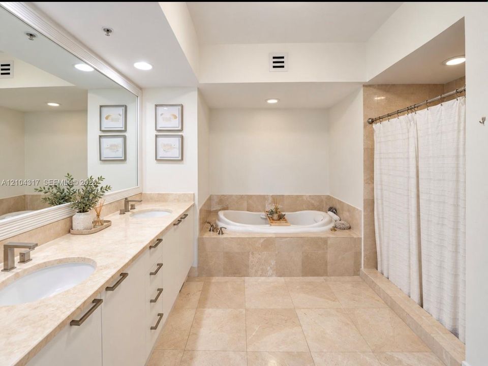 Dual-sink vanity with stone countertop, drop-in soaking tub, stone-tile surround, and full-width wall mirror