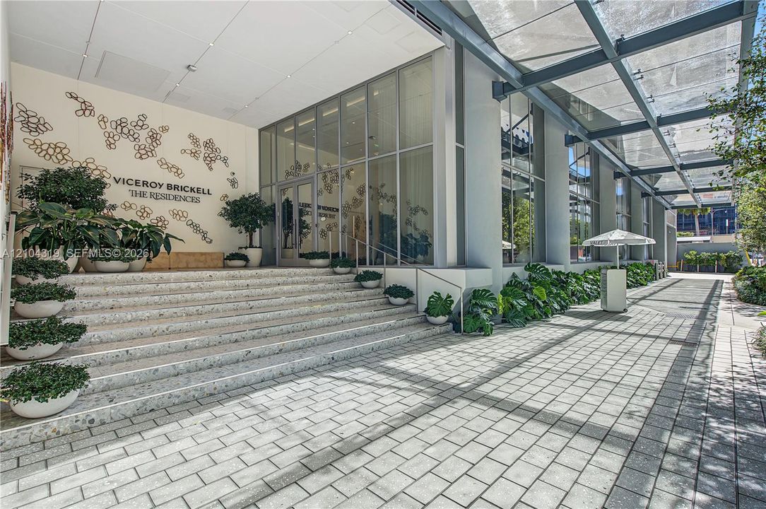 Building exterior featuring a glass curtain wall, expansive paver walkway, overhead steel-frame canopy, and integrated wide-tread stairs with decorative stonework