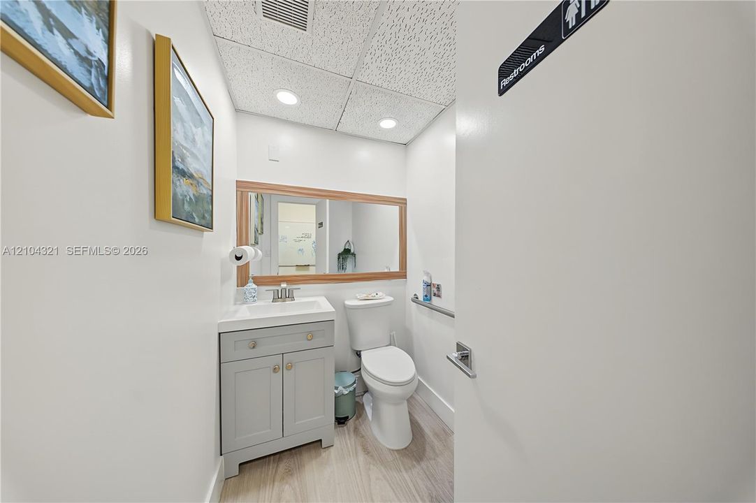 Restroom featuring a grey vanity cabinet with a rectangular sink, wood-finish flooring, a wood-framed wall mirror, and recessed ceiling lighting