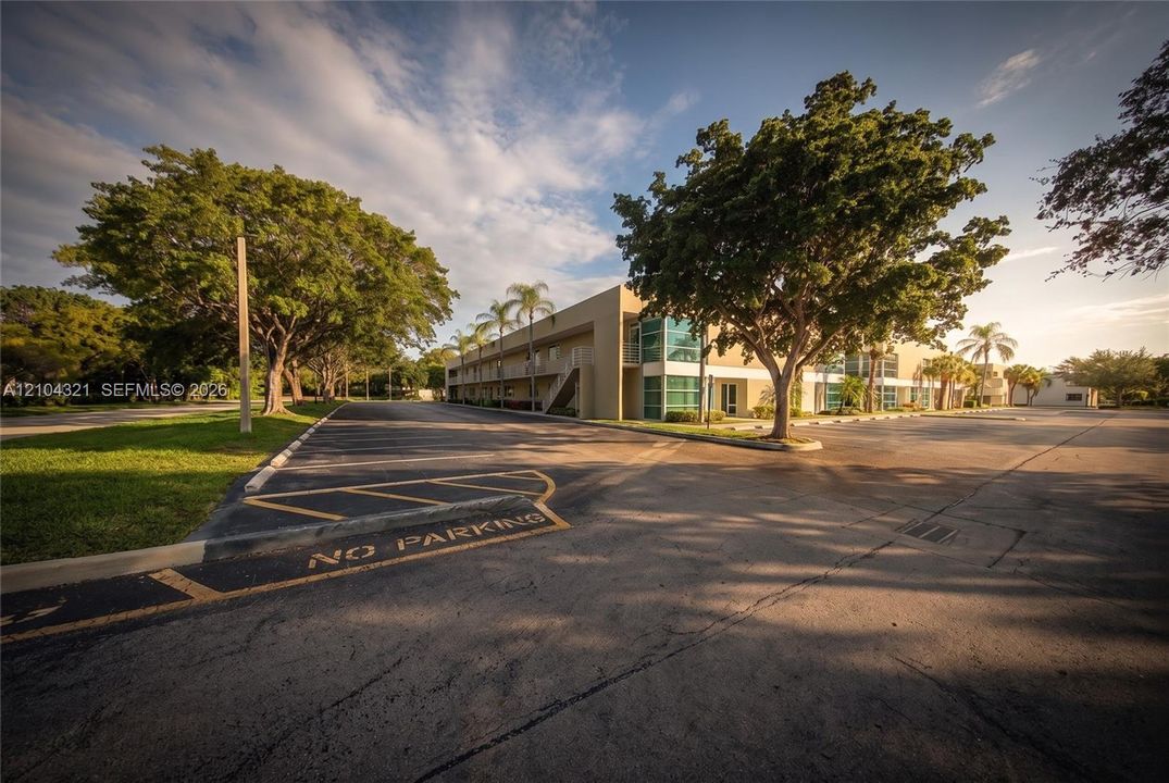 Commercial exterior featuring a multi-story structure with large-pane glass windows, expansive paved parking surface, mature landscaping, and palm trees