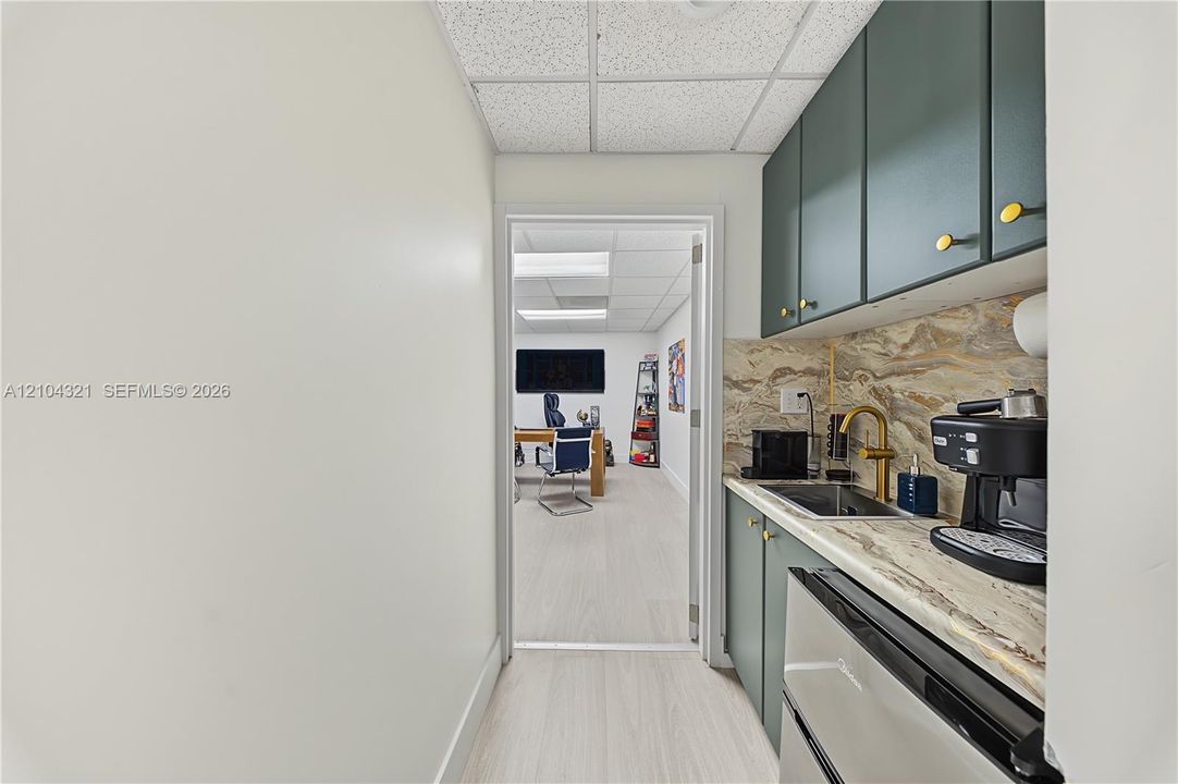 Kitchenette featuring deep green cabinetry, stone-patterned countertops, a stainless steel undermount sink with a brass-finish faucet, and light-toned wood-finish flooring