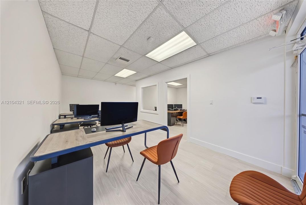 Office area featuring light-toned wood-finish flooring, a suspended acoustic tile ceiling, recessed rectangular lighting fixtures, and a glass partition