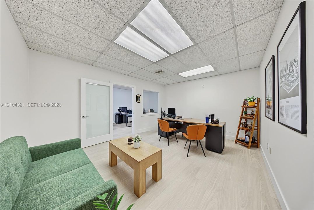 Room featuring light-toned flooring, drop ceiling with integrated lighting panels, white interior doors with glass inserts, and an interior window cutout