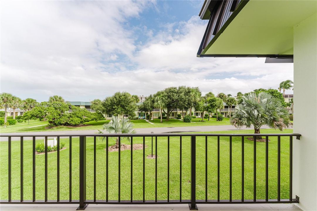 Covered balcony featuring black metal railings and overhead soffit, overlooking a landscaped lawn with mature palm trees