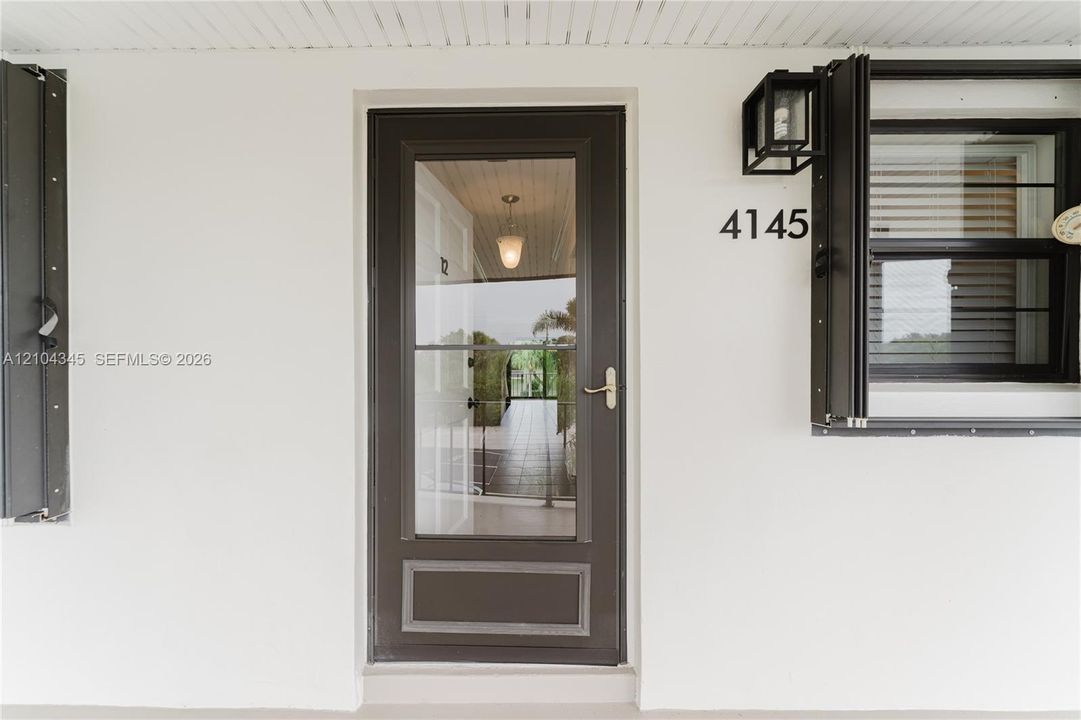 Entryway featuring a dark-toned storm door with glass paneling, metal exterior shutters, a matte black wall-mounted lantern, and high-contrast numerical signage