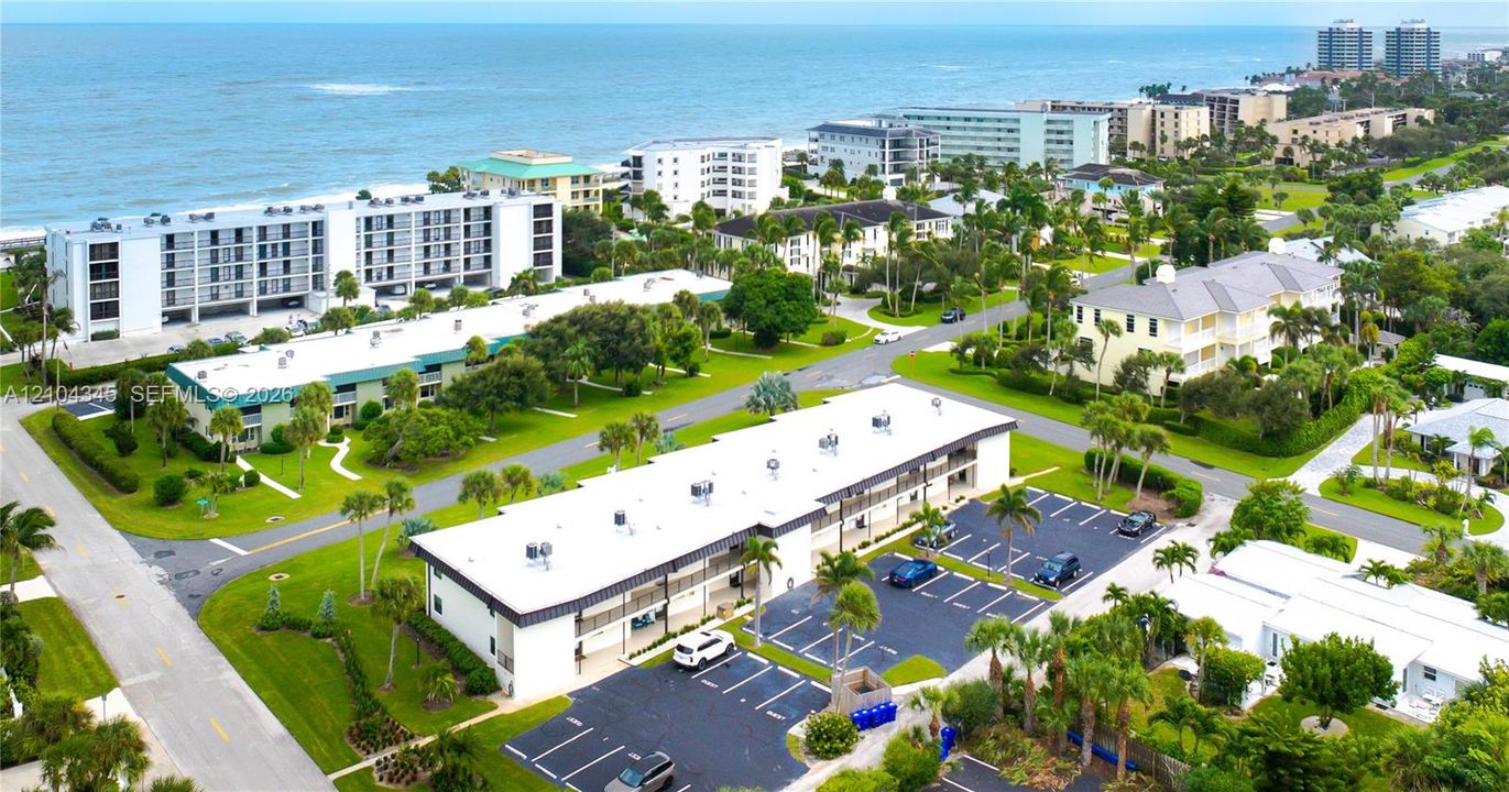 Aerial perspective of multi-unit residential property featuring paved off-street parking, flat roofing, coastal landscaping, and proximity to the oceanfront
