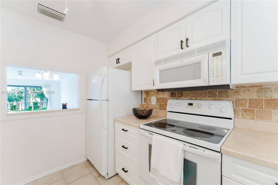 Kitchen featuring white shaker-style cabinetry, a stone-texture backsplash, neutral countertops, white appliances, pass-through to dining/living area.
