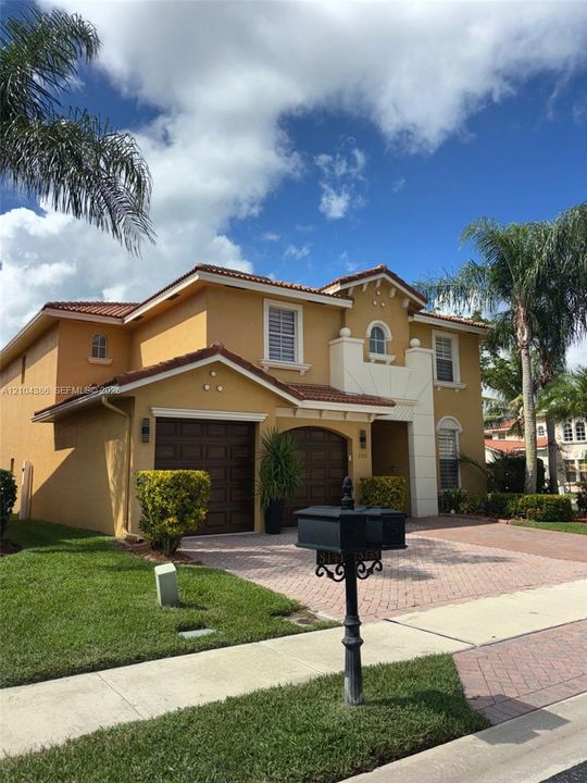 Two-story stucco exterior featuring a herringbone-pattern paver driveway, arched double front entry doors, a multi-car garage, and mature palm landscaping