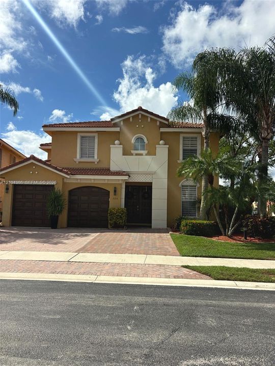 Two-story stucco exterior with a tile roof, paved driveway, mature palm trees, and landscaped gardens with stone edging