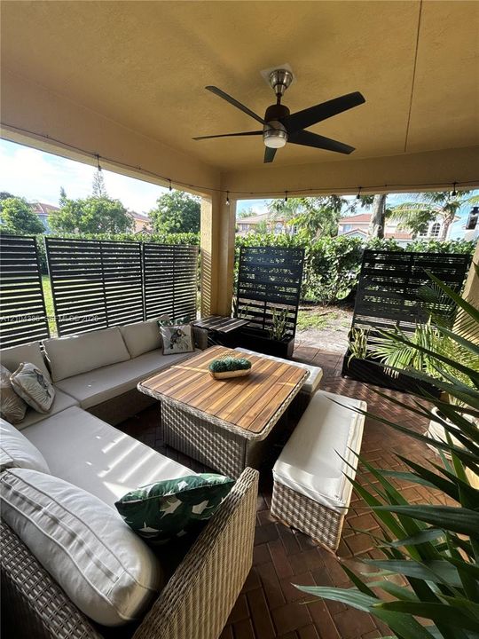 Covered outdoor patio featuring brick paver flooring, a ceiling-mounted fan, and a horizontal slat privacy screen