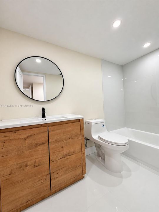 Wood-finish vanity with white countertop, round matte-black framed mirror, matte-black faucet, one-piece dual-flush toilet, and white tile tub surround