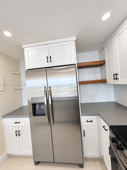 Kitchen featuring white shaker-style cabinetry, matte black hardware, quartz countertops, stainless steel refrigerator, and wood-finish open shelving