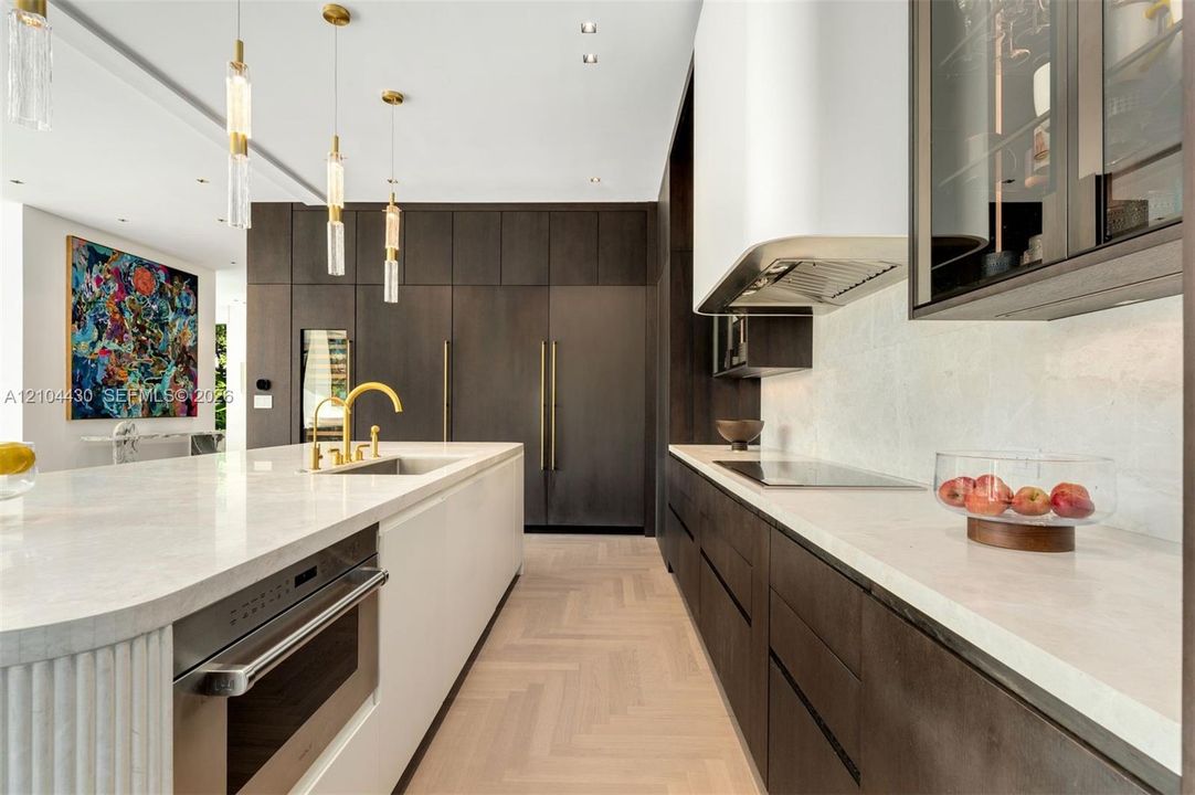 Gourmet kitchen featuring custom dark cabinetry, quartz countertops, a central island with a fluted pillar detail, herring-bone pattern wood-finish flooring, and integrated stainless steel appliances