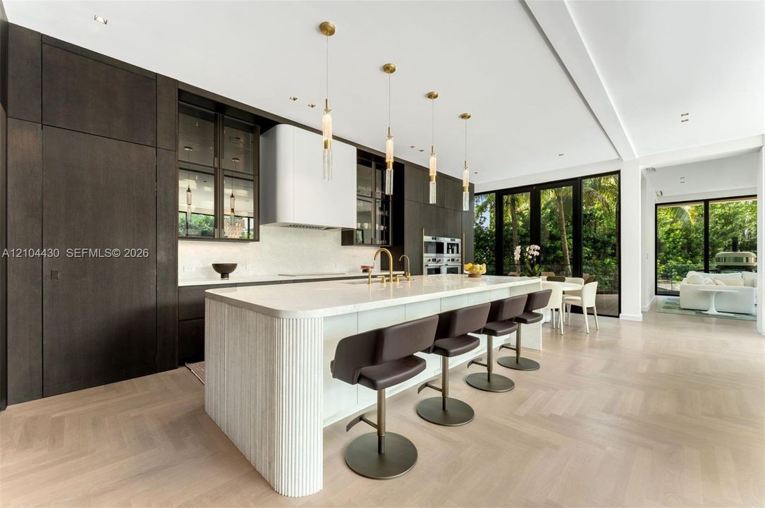 Gourmet kitchen featuring floor-to-ceiling dark wood cabinetry, a large central island with a fluted stone base, herringbone wood-finish flooring, built-in double ovens, and contemporary pendant lighting