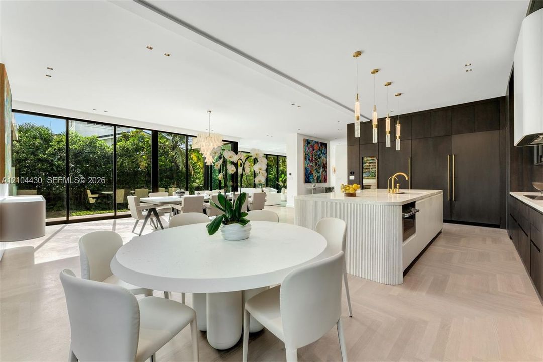 Open-concept kitchen featuring dark floor-to-ceiling cabinetry, a central island with a waterfall countertop, recessed ceiling lighting, multi-pendant illumination, and expansive floor-to-ceiling sliding glass walls
