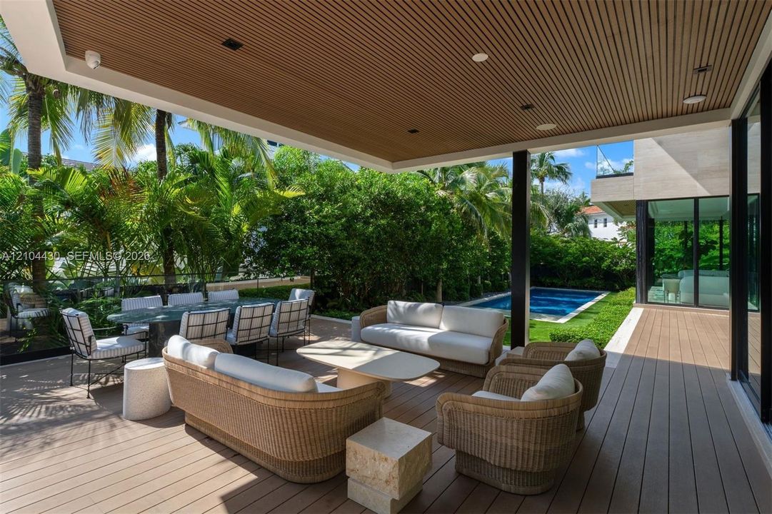 Covered outdoor patio with wood-slatted ceiling, composite decking, recessed lighting, glass railing, and in-ground pool