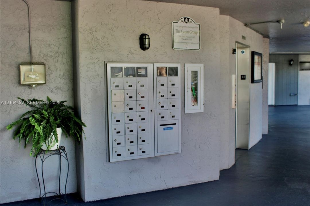 Stucco exterior hallway featuring a centralized metal mail station, integrated fire extinguisher cabinet, and elevator access