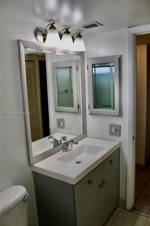 Bathroom vanity featuring a solid-surface countertop with an integrated rectangular sink, three-light brushed-metal vanity fixture, framed wall mirror, and grey built-in storage cabinetry