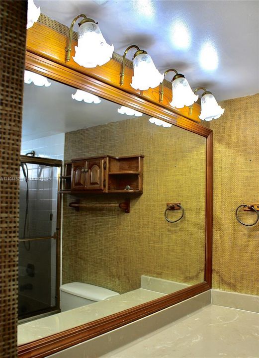 Large, wood-framed wall mirror featuring a four-light fixture with glass shades, wall-mounted cabinetry, and a stone-finish vanity countertop