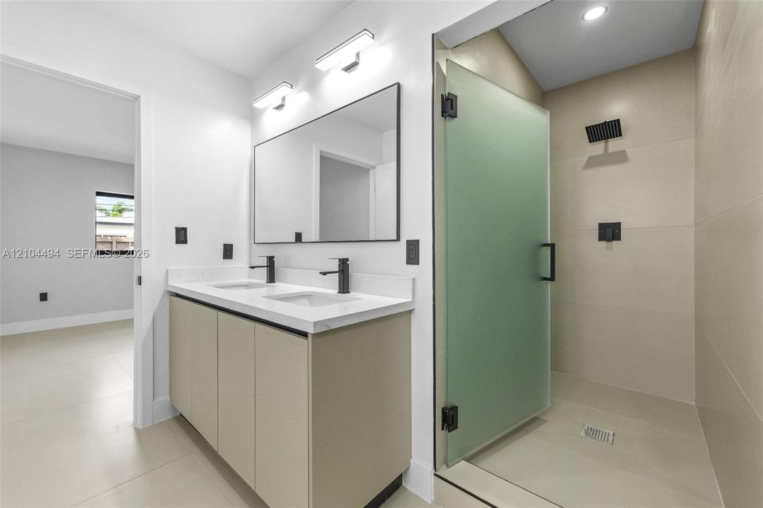 Double vanity with white solid-surface countertop and matte black faucets, paired with a frameless walk-in shower featuring frosted glass, large-format tile surround, and matte black rainfall showerhead