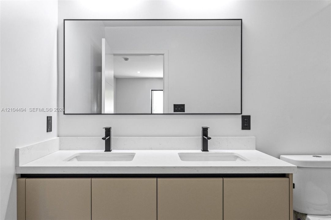 Double vanity featuring solid-surface countertop, dual rectangular undermount sinks, matte black widespread faucets, flat-panel cabinetry, and a large rectangular mirror with a thin black frame