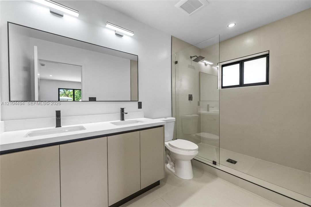 Dual-sink vanity with light-tone cabinetry and white countertop, large wall-mounted mirror, walk-in glass-enclosed shower, and matte black plumbing fixtures